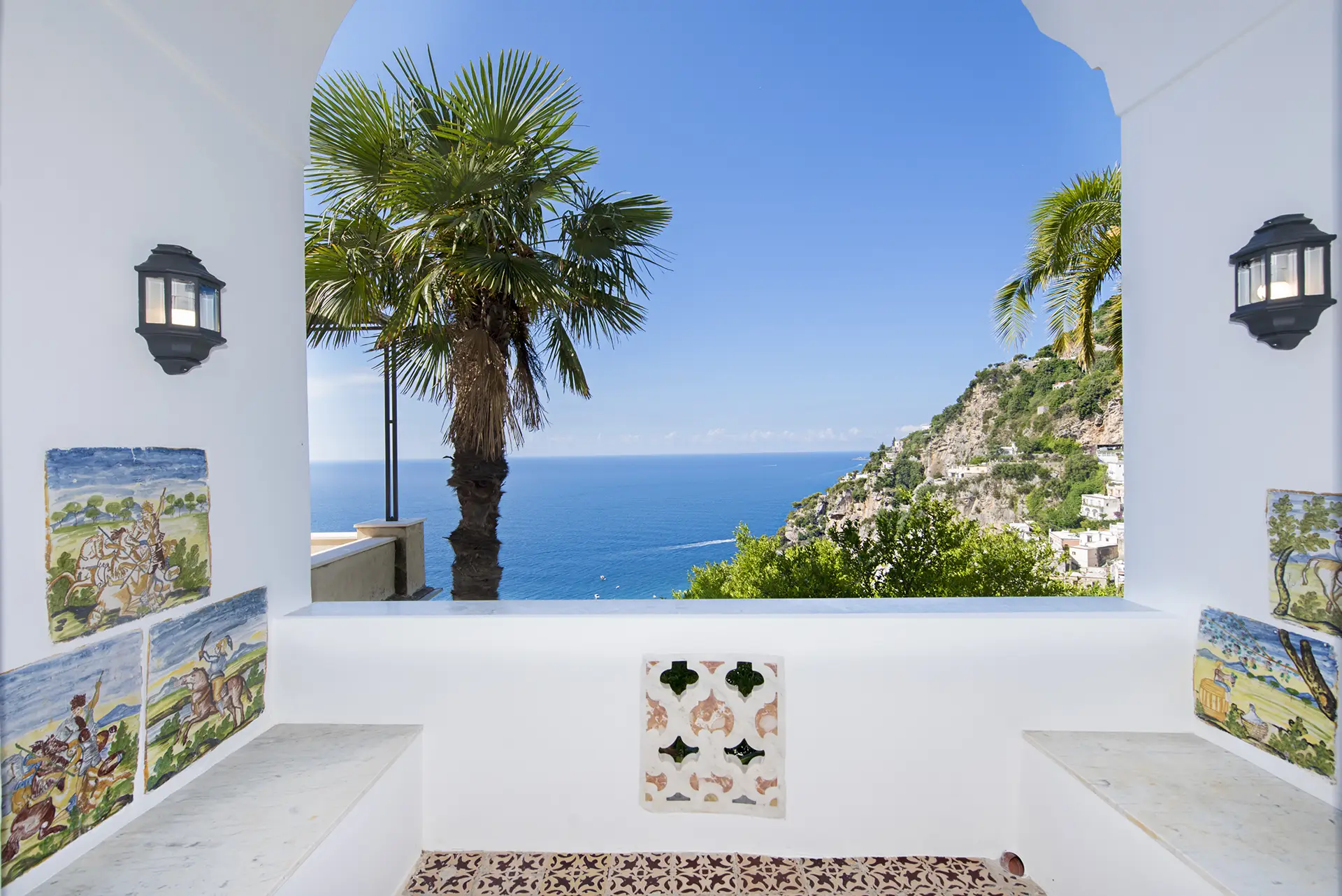 Vista panoramica sul mare e la costa attraverso un arco decorato con piastrelle artistiche e palme.