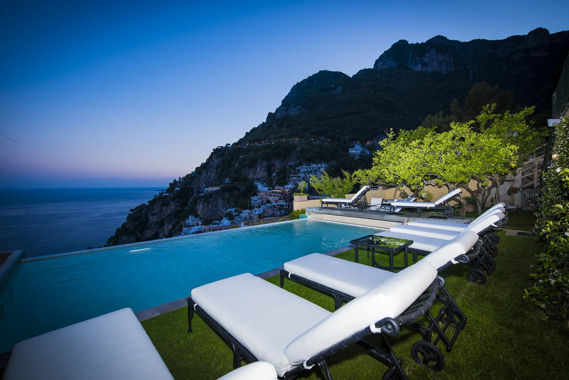 Piscina a sfioro con lettini e vista panoramica sul mare e le montagne al tramonto.