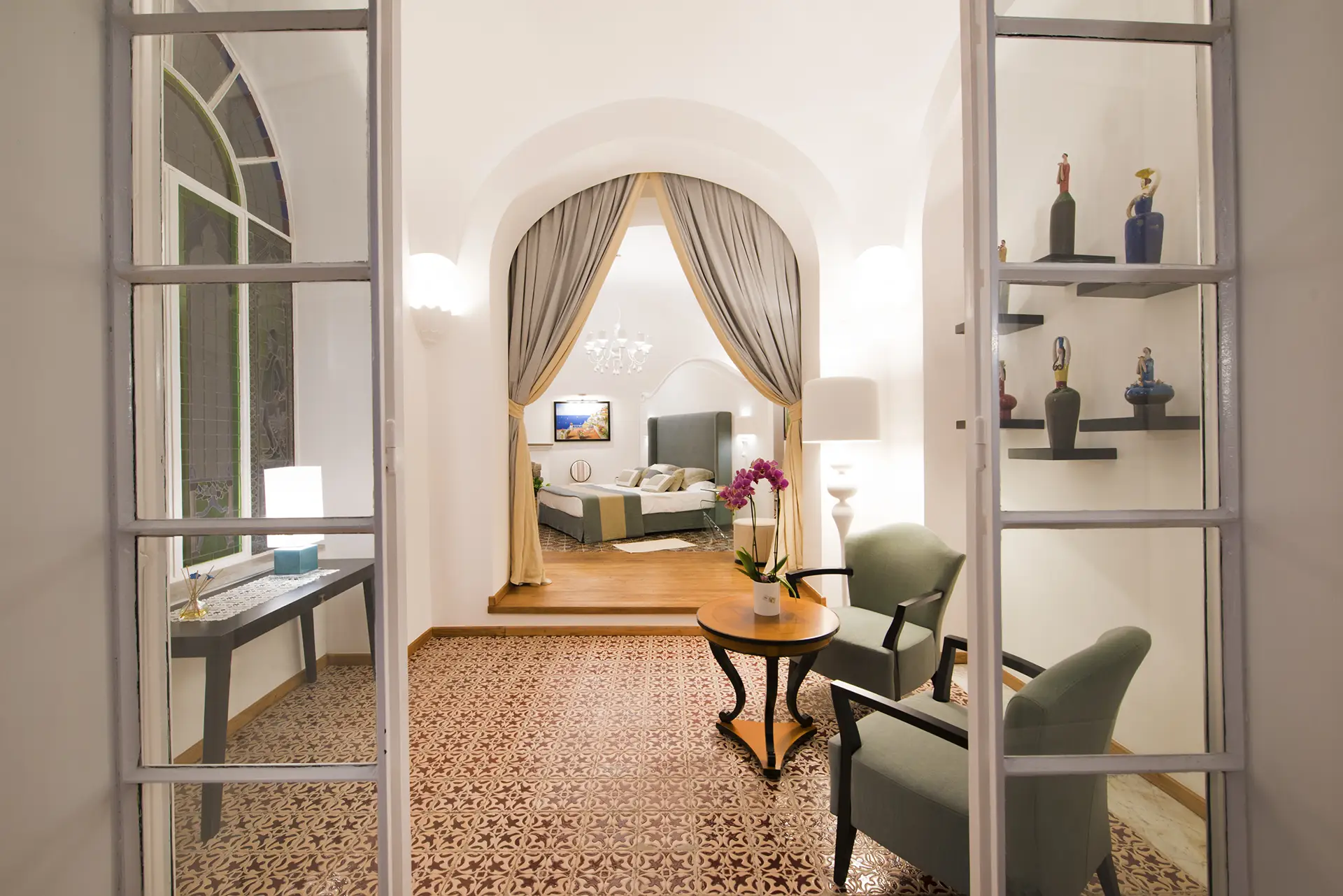 Elegante soggiorno con arredi moderni, lampada, fiori e accesso a una camera da letto accogliente.