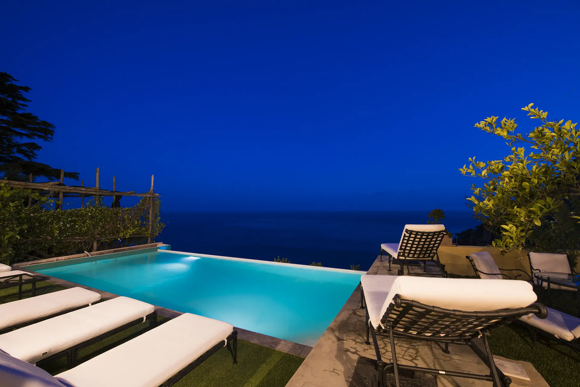 Piscina illuminata di notte con vista sul mare e lettini bianchi.