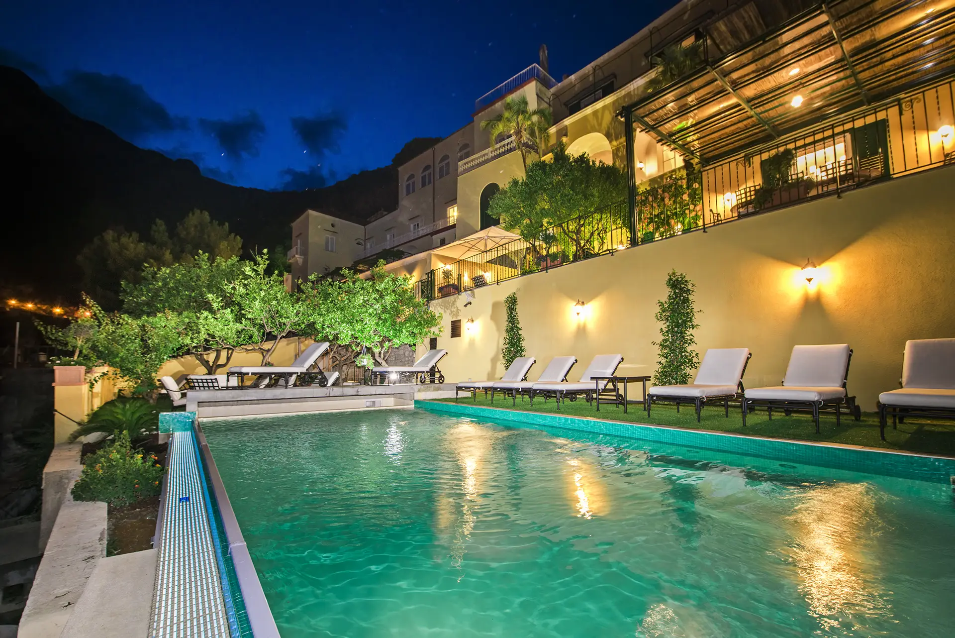 Piscina illuminata di notte con lettini e una villa sullo sfondo.