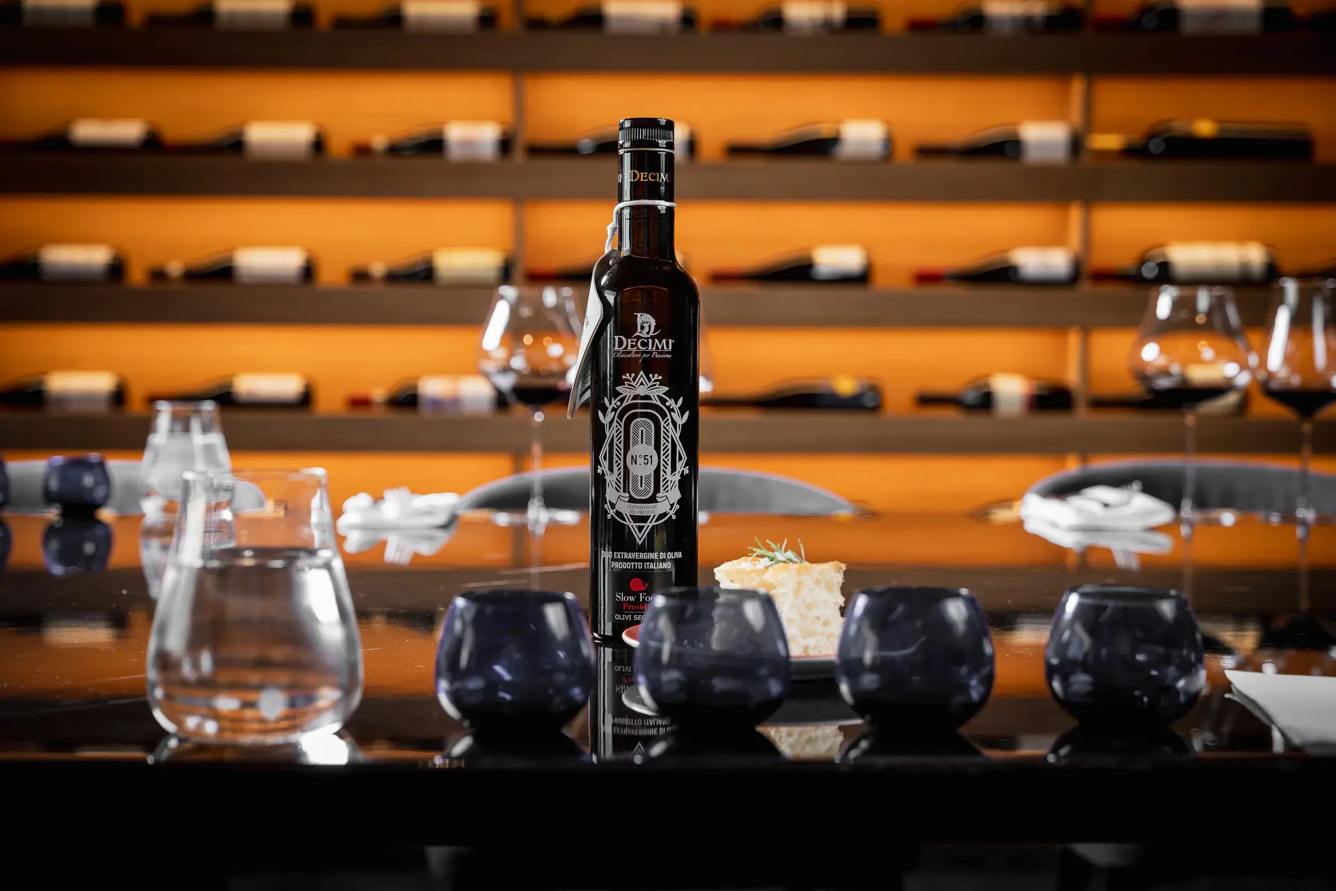 Bottiglia d'olio extravergine di oliva su un tavolo con bicchieri e scaffale di vini sullo sfondo.