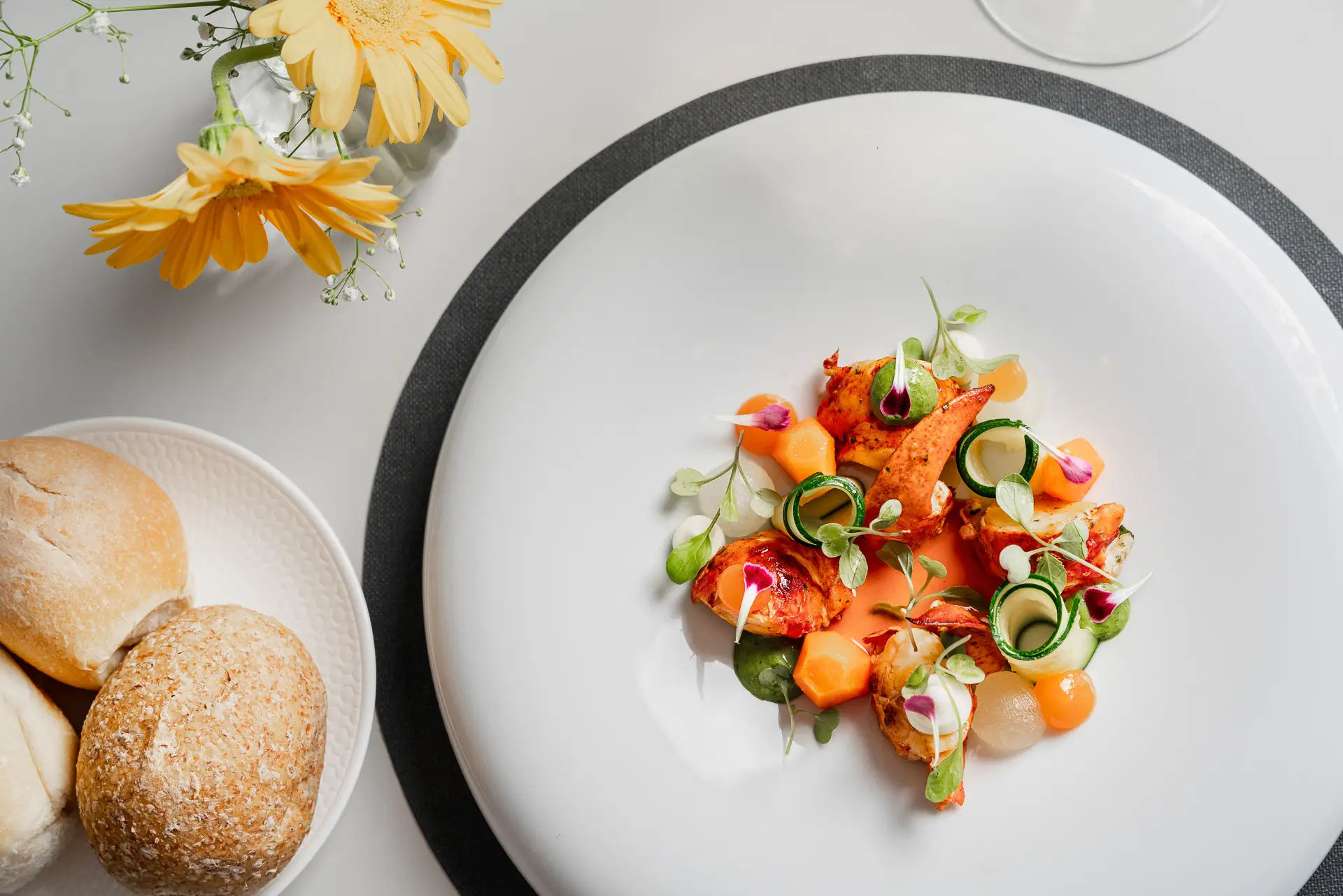 Piatto gourmet con aragosta e verdure decorato elegantemente, accanto a pane e fiori gialli.