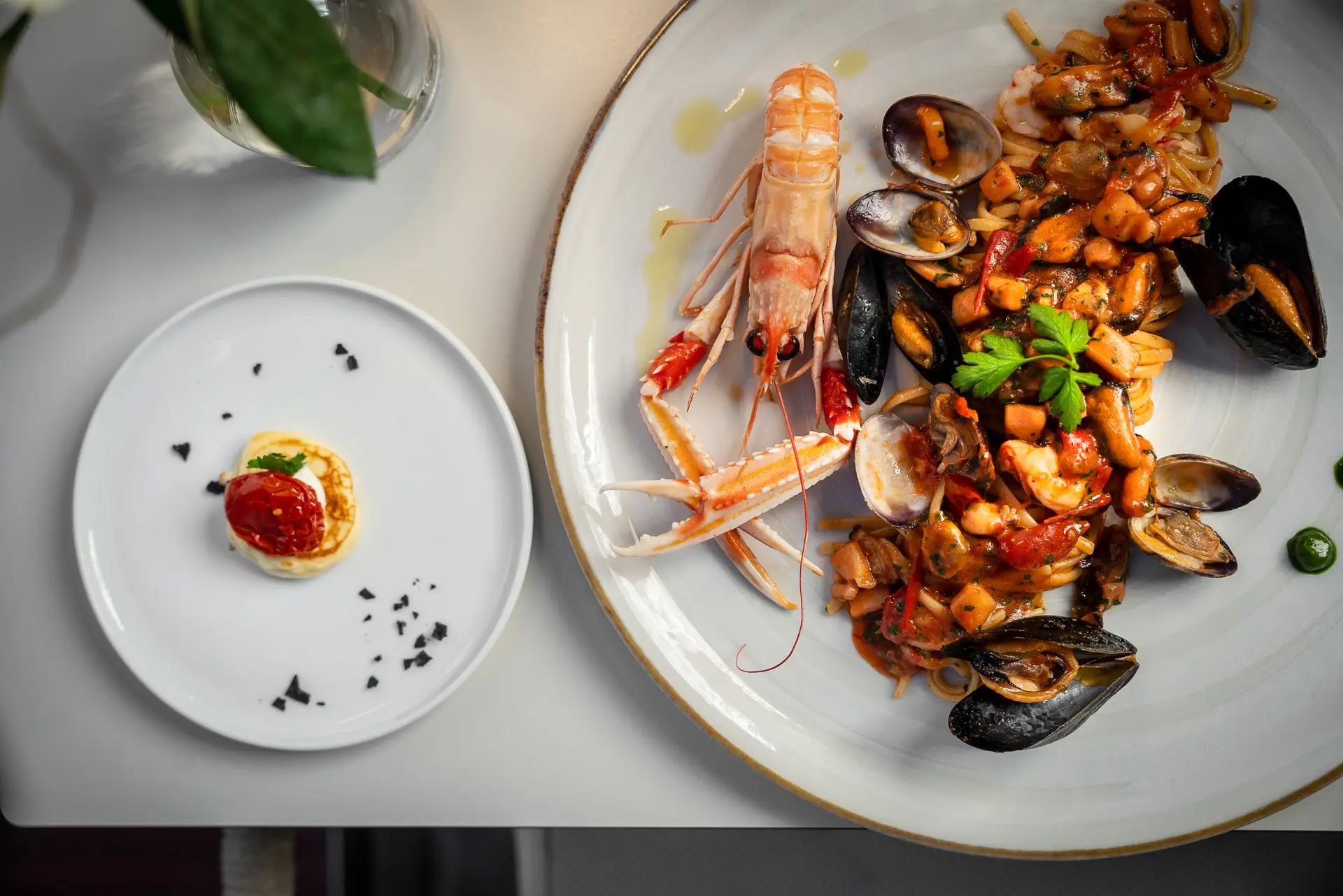 Piatto di spaghetti allo scoglio con frutti di mare e scampo, accanto a un piccolo antipasto su un piatto bianco.