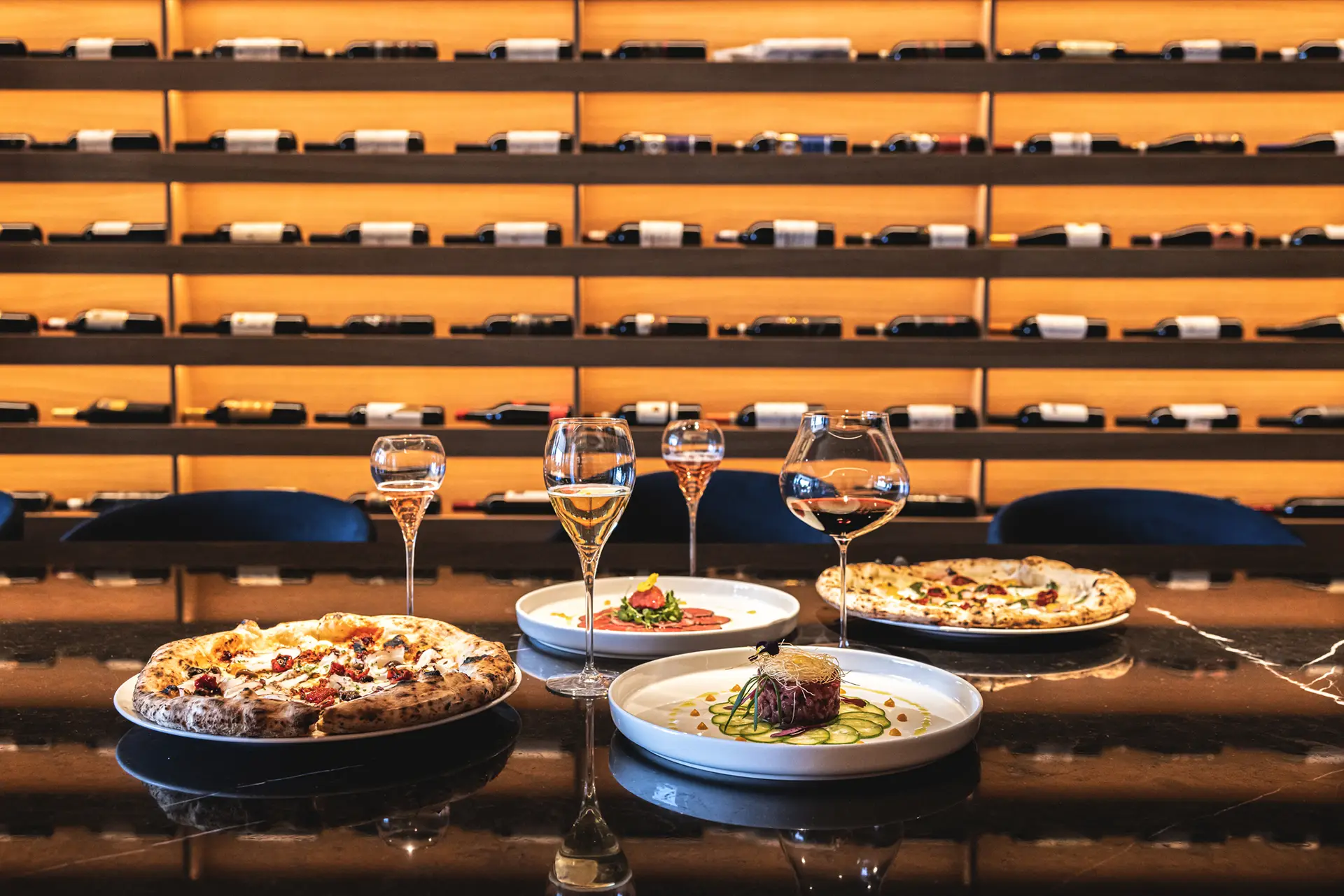 Una tavola apparecchiata con pizze, piatti gourmet e calici di vino, davanti a scaffali di bottiglie illuminati.